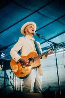 En man i klädd vit kostym och vit hatt, håller i en gitarr och sjunger i en mikrofon.