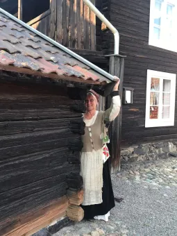 En kvinna iklädd gammeldags kläder, hon håller handen på taket till en mörk timmer byggnad. i bakgrunden en större mörk timmer byggnad med vita fönster.
