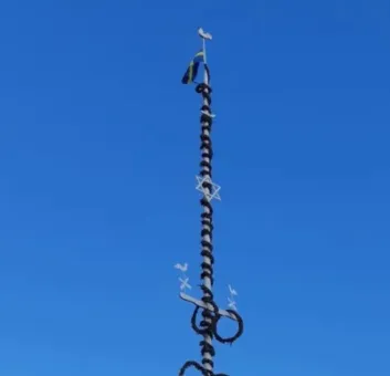 En hög majstång med ringar, dekorationer och en liten flagga syns mot en klarblå himmel..