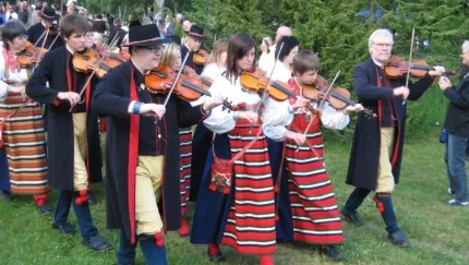 Grupp personer i traditionella folkdräkter spelar fiol utomhus på gräs, med publik och träd i bakgrunden..