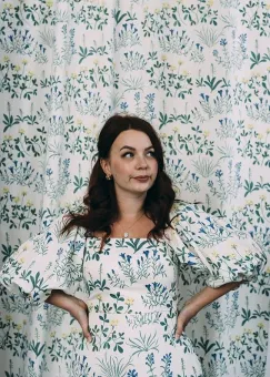 En kvinna med långt mörkt hår, iklädd en vit klänning med blomstermönster, bakom henne tyg i samma mönster som klänningen.