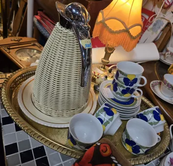 En bricka med en gammal kaffetermos och blommoga kaffekoppar, andra loppis föremål i bakgrunden.