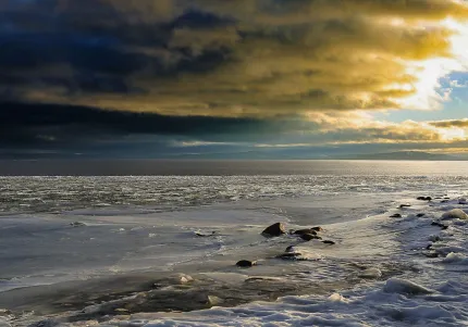 Snötäckt strand med isflak och stenar vid en frusen havsyta, under mörka moln och gyllene solljus vid horisonten..
