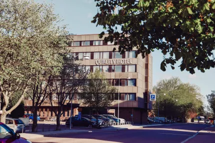 Mid-rise hotel building with a large sign, surrounded by trees, parked cars, and a quiet street in daylight..