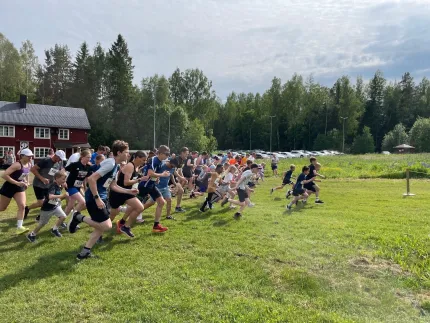 En stor grupp löpare startar samtidigt på en gräsplan nära en röd byggnad, med skog och bilar i bakgrunden..