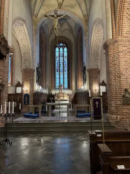 Interiör av en kyrka med tegelpelare, altare och ett stort glasmålat fönster samt kors hängande i taket..
