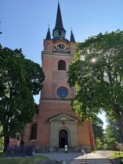 Högt kyrktorn i rött tegel med spira och klocka, inramat av gröna träd mot klarblå himmel och solglimt..