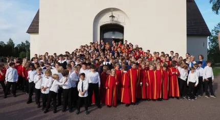 Stor grupp människor i körkläder och vita skjortor samlade utanför en vit kyrkobyggnad i soligt väder..
