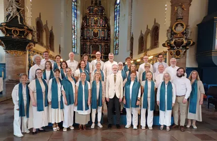 Stor grupp människor i vita kläder med blåa sjalar står uppställda i en kyrka framför altare och glasmålningar..