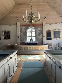 Interiör av ett litet kyrkorum med altare, ljuskrona, bänkar och målningar runt ett centralt fönster..