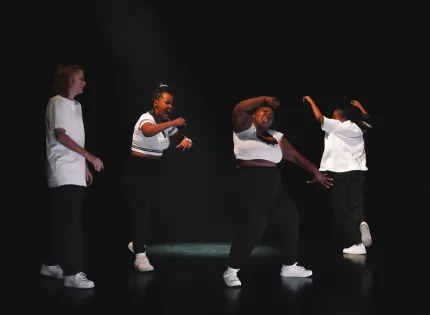 Fyra personer dansar på en mörk scen med spotlight, klädda i vita toppar och svarta byxor med sneakers..