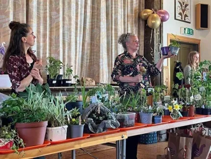 Tre personer bakom ett långt bord fyllt med krukväxter och plantor i en lokal med gardiner och ballonger..