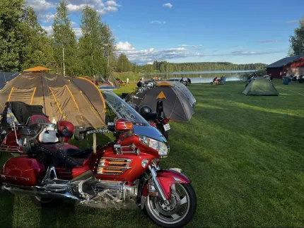 Motorcyklar och tält på en gräsplan, träd och en sjö i bakgrunden.