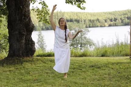 En person i vit klänning dansar barfota på gräs vid en sjö, med stort träd och skog i bakgrunden..