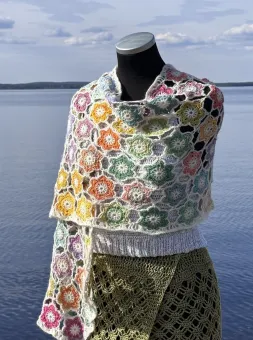 En dockbyst bär en flerfärgad virkad sjal med blomrutor, fotograferad utomhus framför en stilla sjö och molnig himmel..