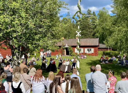 Folk samlas på en gräsmatta och dansar runt en majstång framför en röd träbyggnad omgiven av träd..