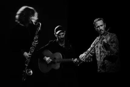 Svartvit bild av tre musiker på mörk scen: en spelar saxofon, en akustisk gitarr och en fiol i sidoljus..