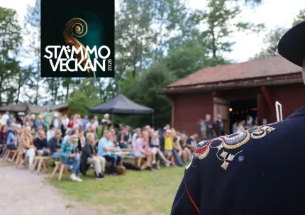 I framkant en axel på en mörk rock som har brodyr, publik som sitter på bänkar, en röd timmerbyggnad, logga Stämmoveckan.