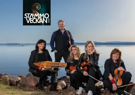 En man och fyra kvinnor alla iklädda svarta kläder och alla håller i ett instrument, mannen står upp och kvinnorna sitter på stenar på en strand, en sjö i bakgrunden, logga Stämmoveckan.