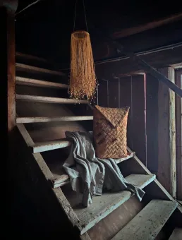 En sliten trätrappa i en mörk stuga med en hängande lampskärm, en vävd kudde och en skjorta på stegen..