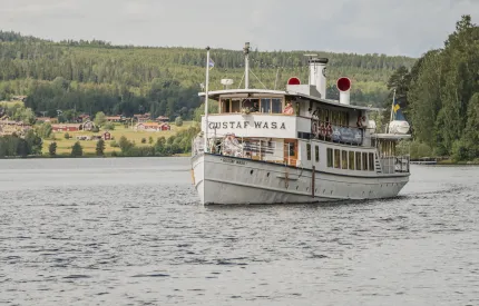 En vit passagerarbåt på en sjö med människor ombord, skogklädda kullar och byhus i bakgrunden..