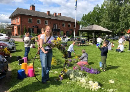 En kvinna i långt ljust hår iklädd vit t-shirt och jeans som placerar små blomsterbuketter på en ring, barn och vuxna på en gräsmatta som också binder kransar, en röd byggnad i bakgrunden.