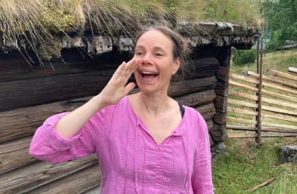 En person i rosa blus står framför en gammal timmerstuga med grästak och ett trästaket i bakgrunden..