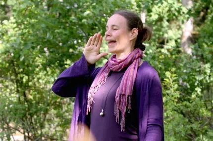 En person står utomhus bland gröna träd, med hand uppsträckt mot kameran och lila kofta samt scarf..