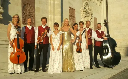 Grupp musiker i festkläder står vid en stenbyggnad i sol, med cello, fioler och kontrabas..