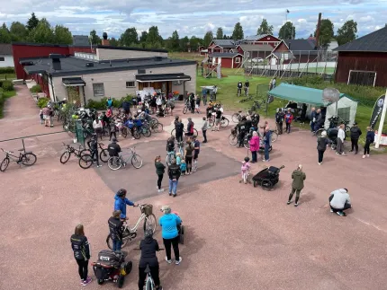 Många personer och cyklar samlade på en gårdsplan utanför byggnader, med tält och molnig himmel i bakgrunden..