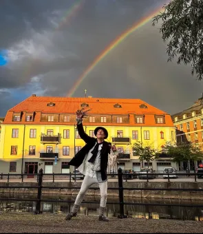 En person poserar vid en kanal framför ett gult hus med rött tak, medan en regnbåge syns på mörk himmel..