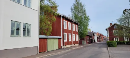 En stilla gata med röda trähus och vita knutar, ett vitt hus i förgrunden samt gröna träd under klar himmel..