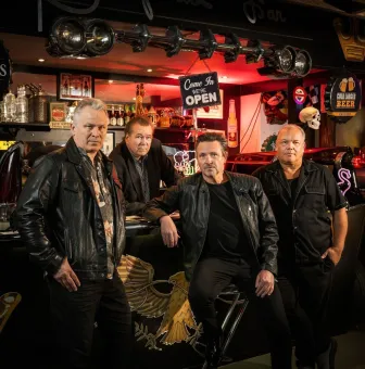 Fyra män i läderkläder poserar vid en bar med flaskor, neonskyltar och röd belysning i bakgrunden..