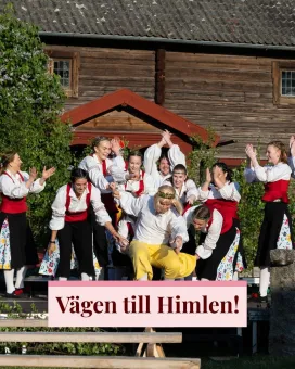 Grupp i folkdräkter uppträder på utomhusscen framför träbyggnad, med skylttexten "Vägen till Himlen!"..