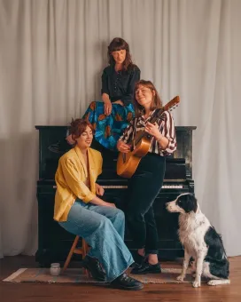 Tre kvinnor vid ett piano, en av dem sitter uppe på pianoten sitter på en stol brevid och den tredje står och håller i en gitarr, en svartvit huns sitter på golvet och tittar.