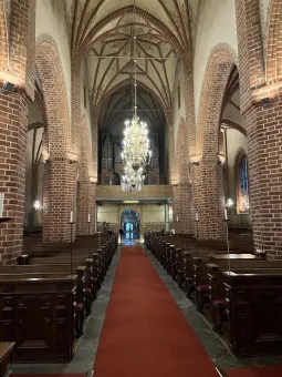 Interiör av en kyrka med tegelvalv, bänkrader och en röd mittgång som leder mot orgeln och en stor ljuskrona..