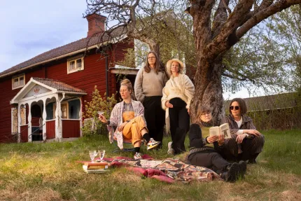 Fem personer sitter och står vid ett stort träd på gräsmatta med filt, böcker och glas framför ett rött hus..