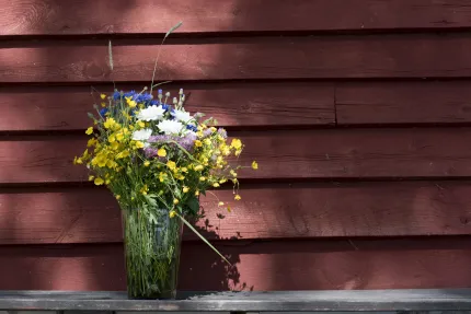 En bukett vildblommor i en glasvas står på en grå bänk framför en röd trävägg i solljus..