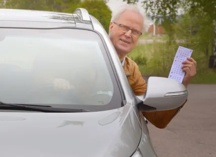 En man i grått hår och glasögon, sticker ut huvudet från bilrutan på en bil, håller en bingobricka i handen.