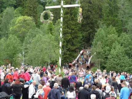 En midsommarstång, en stor grupp besökare som har samlats runt stången.