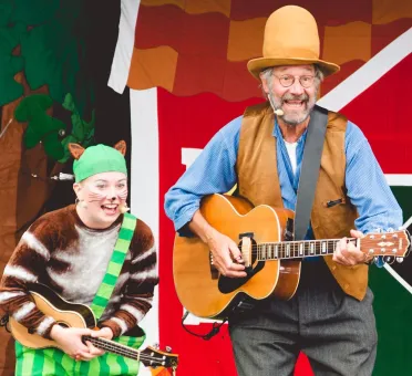En kvinna utklädd till katten Findus, en man i stor hög hatt väst som spelar på en gitarr, teaterkuliss i form av en röd stuga i bakgrunden.