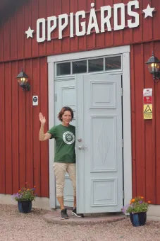 En kvinna med kort hår,  iklädd grön t-shirt och beiga byxor, hon står i en dörröppning och vinkar, en röd träbyggnad med Oppigårds över dörren.