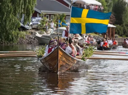 Träbåt på vatten med människor i folkdräkter och blomsterkransar; en stor svensk flagga vajar i fören..