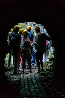 En grupp personer med gula hjälmar står i en grotta och tittar ut mot en ljus, stenig skogsmiljö..