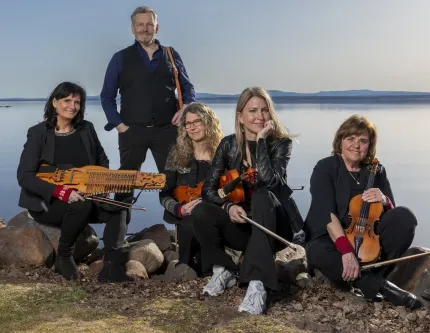 Fyra kvinnor i mörka kläder som sitter på stenar på en strand, tre av dem håller i varsin fiol och den fjärde håller i en nyckelharpa, bakom dem står en man i mörka kläder, i bakgrunden vatten och blånande berg.
