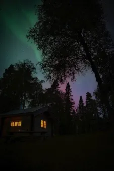 En bild tagen på natten, ljusfenomen på himlen, träd, en stuga där man ser ljus från fönstren.