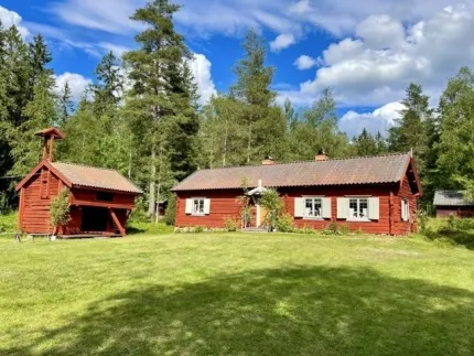 En röd timmerstuga och ett rött härbre, gårdstun, skog i bakgrunden.