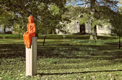 En staty föreställande en sittande kvinna, park och en vit byggnad i bakgrunden.