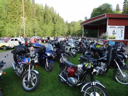 En grupp motorcyklar i framkant, bakom dem veteranbilar och en röd folkparksscen.