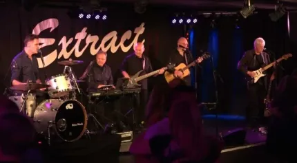 Fem män på en scen, tre av denspelar gitarr, en keyboard och en trummor, Extract med guldbokstäver.